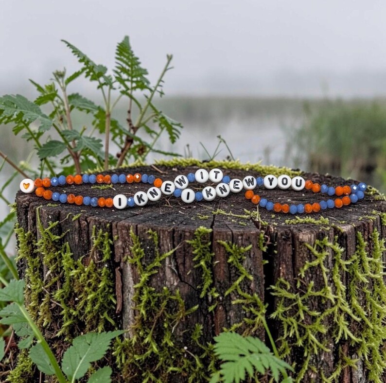 May include: A beaded bracelet featuring alternating orange and blue faceted beads, interspersed with white round beads displaying the black text I KNEW IT I KNEW YOU, and a single small orange heart bead at the end of the beaded strand.