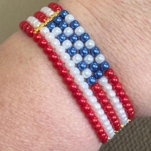 Op de afbeelding: Een kralenarmband met een ontwerp dat doet denken aan de Amerikaanse vlag. De armband bestaat uit rode, witte en blauwe kralen, met goudkleurige accenten. De kralen zijn in rijen gerangschikt om de strepen en sterren van de vlag na te bootsen.