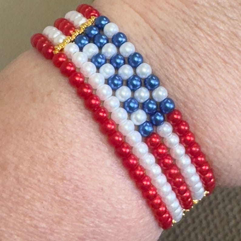 Op de afbeelding: Een kralenarmband met een ontwerp dat doet denken aan de Amerikaanse vlag. De armband bestaat uit rode, witte en blauwe kralen, met goudkleurige accenten. De kralen zijn in rijen gerangschikt om de strepen en sterren van de vlag na te bootsen.