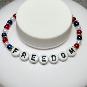 A patriotic bracelet with the word "FREEDOM" spelled out in white letter beads. The bracelet features red, white, and blue beads, creating a festive and colorful accessory. The bracelet is a stretch style.