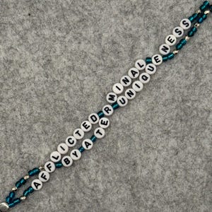 May include: A beaded lanyard with the words "AFFECTED BY A TERMINAL UNIQUENESS" spelled out in white letter beads. The beads are strung on teal-colored string with silver accents. The lanyard is on a gray background.