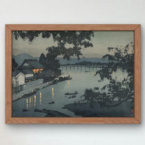 以下が含まれることがあります： 橋と家々が川岸に沿って建ち並ぶ、夜の川の風景を描いた、額装された日本の木版画。版画は青、グレー、白の色合いで、黄色い光が差し込んでいます。