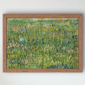 Op de afbeelding: Een ingelijst schilderij met een dicht veld van hoog groen gras, afgewisseld met kleine gele, roze en blauwe wilde bloemen. Het kunstwerk bevat zichtbare, getextureerde penseelstreken in verschillende tinten groen, wat een levendig weidelandschap vormt in een eenvoudige, lichtbruine houten lijst.