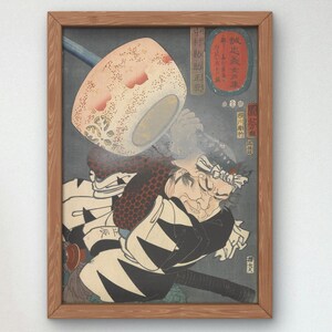 Könnte beinhalten: Ein gerahmter japanischer Holzschnitt, der einen Samurai-Krieger zeigt, der ein großes, rundes, weißes und rotes Objekt hält. Der Krieger trägt eine schwarz-weiß gemusterte Robe und ein weißes Stirnband. Der Druck ist vor einem dunkelblauen Hintergrund platziert und enthält japanischen Text.