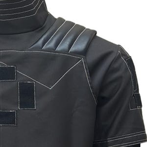 May include: Black short-sleeved garment with white stitching details. Features a high collar and padded shoulder accents. Includes rectangular patches with velcro closures. The design is modern and sleek.