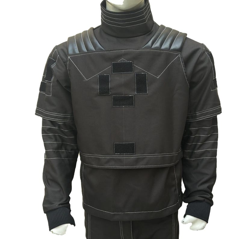 May include: A dark gray jacket with a high, ribbed collar and white stitching details. The jacket features black patches and shoulder accents. The sleeves have a layered design with white stitching, and the cuffs are ribbed.
