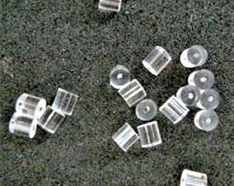 May include: A close-up of a group of clear plastic earring backs. The backs are small and round, with a slight indentation in the center. They are scattered on a gray surface.
