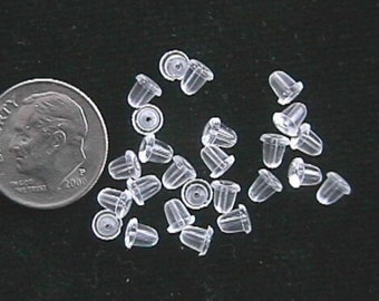 May include: A close-up of a pile of clear plastic earring backs. The backs are small and round, with a slight indentation in the center. They are designed to be used with stud earrings.