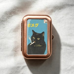 May include: A rectangular copper-toned metal case featuring a graphic insert of a black cat with yellow eyes smoking a cigarette against a light blue background. The graphic includes yellow Japanese text at the top and smaller white text below the cat.