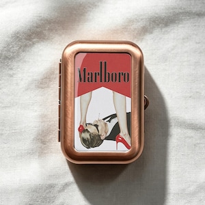 May include: A rectangular copper-toned metal cigarette case featuring a central graphic insert. The image displays the text Marlboro above a stylised illustration of a person in a suit and sunglasses with red high heels positioned above them.