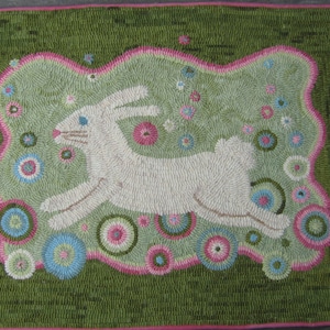 May include: Hand-tufted rug featuring a white rabbit leaping across a light green field. The rabbit is surrounded by colorful floral accents in pink, blue, and green. The rug has a dark green border with a pink trim.