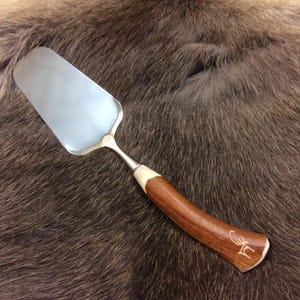 May include: A silver cake server with a polished blade and a handle made of dark brown wood. The handle has a curved design with a light-coloured accent and a carved initial. The server is set against a brown fur background.