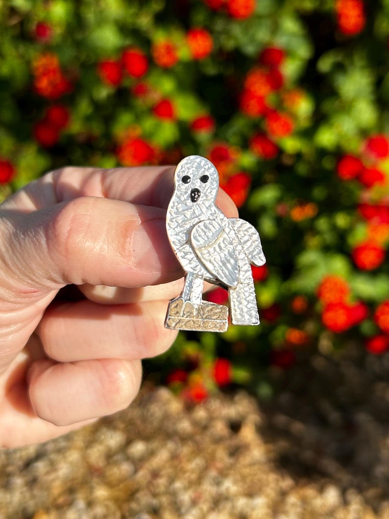 May include: A silver owl brooch with black eyes, perched on a textured base. The owl has a detailed, textured surface.