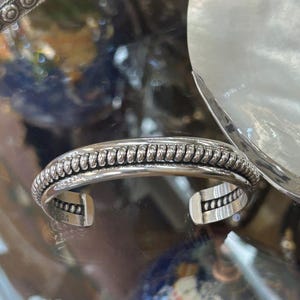 May include: A silver cuff bracelet with a textured, rope-like design along the top edge. The open-ended bracelet has a polished finish and is displayed on a reflective surface. The word "STER" is stamped on the inside of the bracelet.