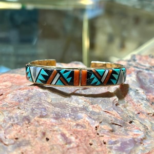 May include: A gold-toned cuff bracelet with a geometric design. The bracelet features turquoise, black, white, and orange inlaid stones. The design is a series of triangles and rectangular shapes. The bracelet is displayed on a textured, reddish-brown surface.