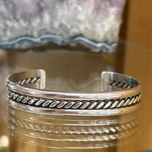 May include: A silver cuff bracelet featuring a central twisted rope design flanked by smooth, polished silver bands. The open-ended bracelet has a reflective metallic finish and a consistent width throughout its circular, rigid structure.