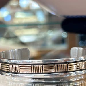 Puede incluir: Brazalete de plata con una banda decorativa de patrones rectangulares dorados. El brazalete tiene un diseño sencillo y abierto y se muestra sobre una superficie reflectante.