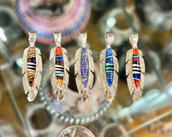 May include: Five silver feather pendants featuring inlaid stone designs. Each pendant displays a unique pattern of colored stones, including orange, blue, purple, black, and white segments. A United States quarter coin is visible for scale.