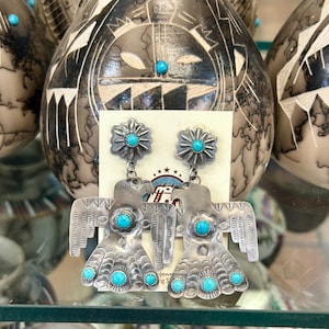 May include: A pair of silver earrings featuring turquoise stones. The earrings are shaped like a bird with outstretched wings. The turquoise stones are set in a silver flower design.