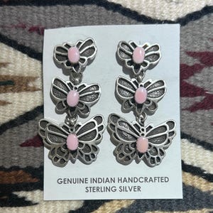 May include: A pair of sterling silver dangle earrings with a butterfly design. Each earring features three butterfly shapes with pink center stones. The card reads "GENUINE INDIAN HANDCRAFTED STERLING SILVER."