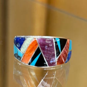 May include: A wide band ring featuring a multi-colored inlay design. The surface is composed of geometric segments of lapis lazuli, turquoise, orange spiny oyster, purple spiny oyster, and black onyx, all separated by thin silver metal dividers.
