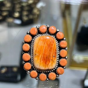 May include: A silver ring featuring a large, rectangular, orange-streaked stone at the center, surrounded by a border of fourteen small, round, peach-colored cabochons. The central stone is framed by a twisted silver rope detail.