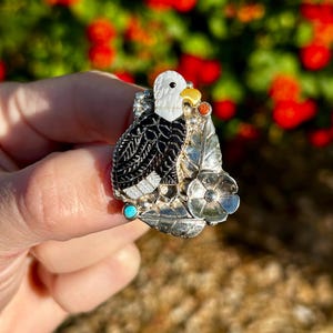 May include: A silver ring featuring a black and white eagle design, accented with turquoise and red stones. The eagle is perched on a branch with leaves and a flower.