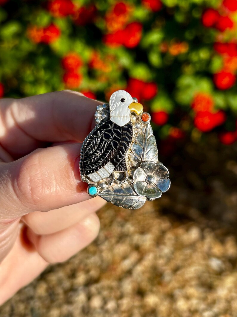 May include: A silver ring featuring a black and white eagle design, accented with turquoise and red stones. The eagle is perched on a branch with leaves and a flower.