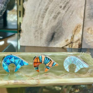 May include: Three silver pendants in a bear claw design, inlaid with turquoise, orange, and opal-like stones. The pendants are displayed on a green, marble-like surface, showcasing the intricate details of the jewelry.