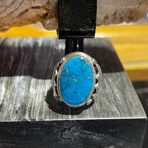 May include: Silver ring featuring a vibrant turquoise oval stone. The stone is set in a silver band with decorative detailing. The ring is displayed on a dark, textured surface.