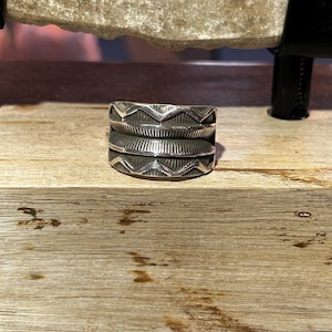 May include: A wide silver band ring featuring a geometric pattern of triangles and lines. The ring is crafted from sterling silver and has a polished finish.