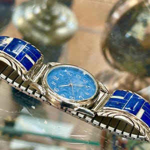 Op de afbeelding: Een zilverkleurig horloge met een blauwe wijzerplaat en bijpassende blauw-witte inleg op de band. Het horloge heeft een ronde wijzerplaat met zilveren wijzers en uurmarkeringen. De band is gemaakt van zilveren schakels.