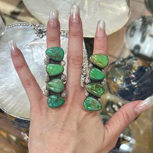 Puede incluir: Dos anillos de plata con cinco piedras ovaladas de turquesa verde brillante cada uno. Los anillos tienen un diseño floral decorativo entre las piedras. Los anillos se muestran en una mano.