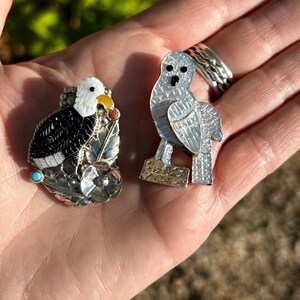 May include: Two silver and gemstone bird pins. The first pin is a black and white eagle with turquoise and yellow accents. The second pin is a white owl with black eyes.