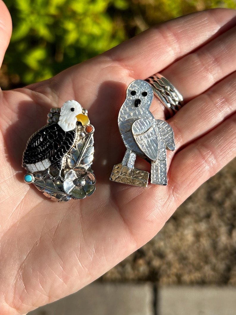 May include: Two silver and gemstone bird pins. The first pin is a black and white eagle with turquoise and yellow accents. The second pin is a white owl with black eyes.