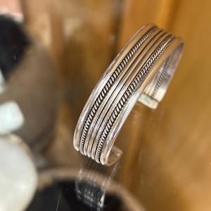 May include: A silver cuff bracelet featuring a polished finish with three distinct horizontal bands. The design includes two outer twisted rope-style textures flanking a smooth central bar, creating a layered appearance with a reflective metallic surface.