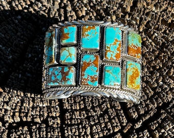 May include: A silver and turquoise bracelet with a geometric design. The bracelet is made of sterling silver and features a row of turquoise stones set in a square pattern.
