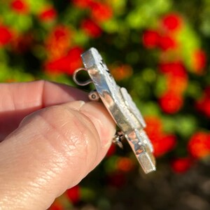 May include: A silver brooch with a textured surface and a pin on the back. The brooch is shaped like a butterfly with a white centre and black accents.