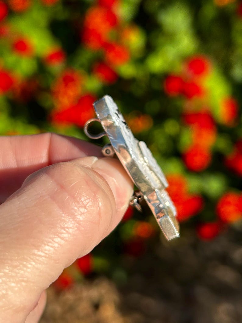 May include: A silver brooch with a textured surface and a pin on the back. The brooch is shaped like a butterfly with a white centre and black accents.
