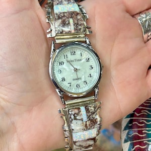 May include: A silver quartz watch with a white face and black numerals. The watch band features a mosaic design with brown and white stone inlays. The watch is held in a hand.