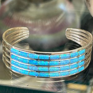 May include: Silver cuff bracelet with four rows of bright blue rectangular inlays. The bracelet has a simple, open design with multiple parallel bands. The blue inlays have a shimmering, opalescent quality.