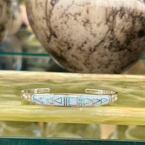 Puede incluir: Una pulsera de plata con incrustaciones de ópalo rectangular con diseños geométricos. El ópalo tiene una combinación de colores azul claro y blanco. La pulsera se exhibe sobre una superficie verde.