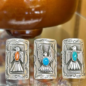 May include: Three rectangular silver rings featuring embossed bird motifs. The left ring has an oval orange stone, the centre ring has an oval turquoise stone with wings spread, and the right ring has an oval turquoise stone at the centre.