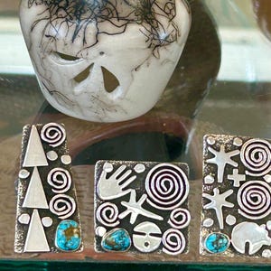 May include: Three silver-toned rectangular pendants with turquoise stones and various designs. The designs include triangles, spirals, handprints, stars, and a bear. A decorative ceramic piece is in the background.