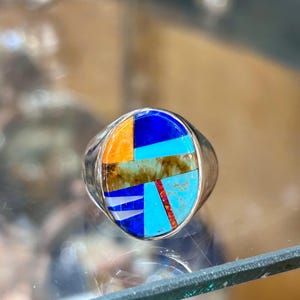 May include: A silver ring with a colorful inlaid design. The ring's face features a mosaic of geometric shapes in orange, blue, turquoise, and red. The ring has a polished, rounded band.