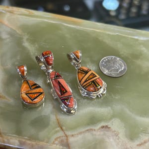 May include: Four silver pendants with inlaid orange and black geometric designs. The pendants have different shapes, including teardrop and oval. A US dime is next to the pendants for size comparison. The pendants are displayed on a green surface.