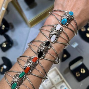 May include: A stack of six silver cuff bracelets, each featuring a central oval gemstone. The stones include turquoise, white opal, black onyx, red coral, green malachite, and a second turquoise, all set in decorative silver metalwork.
