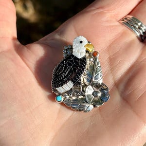 May include: A silver pendant featuring a detailed depiction of an eagle with black, white, and yellow accents. The eagle is perched on a leaf with a turquoise stone and a flower.