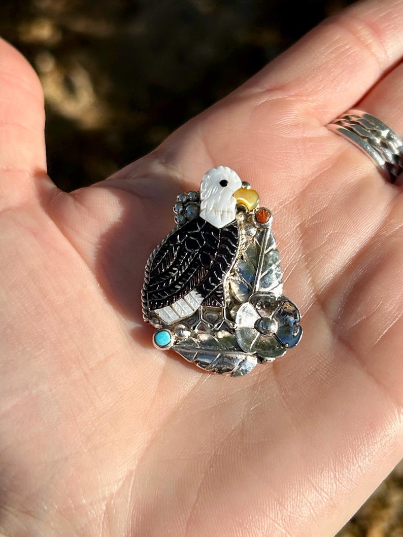 May include: A silver pendant featuring a detailed depiction of an eagle with black, white, and yellow accents. The eagle is perched on a leaf with a turquoise stone and a flower.