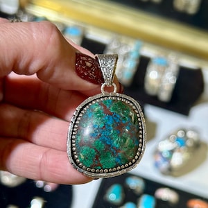 Puede incluir: Un colgante de plata con una piedra preciosa verde y azul engastada en un engaste de plata. El colgante tiene un borde de cuentas y una anilla texturizada.